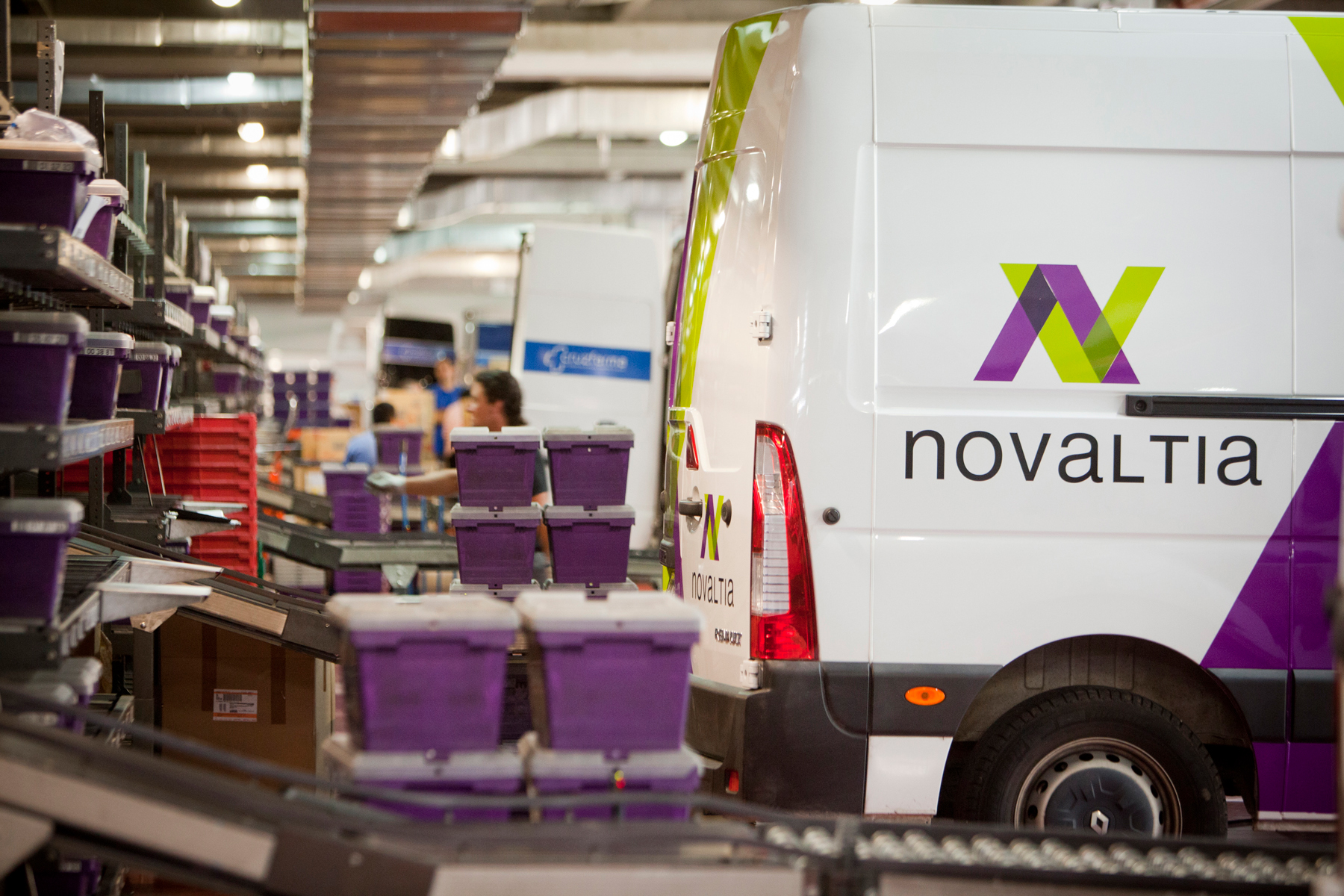 Furgoneta de reparto Novaltia en un centro logístico farmacéutico con contenedores y sistemas de clasificación de pedidos en funcionamiento.