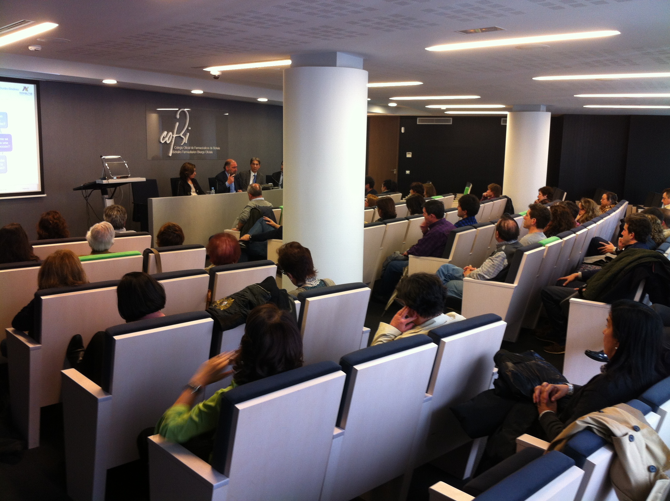 Asistentes sentados en una sala de conferencias mientras varios ponentes intervienen desde la mesa principal.