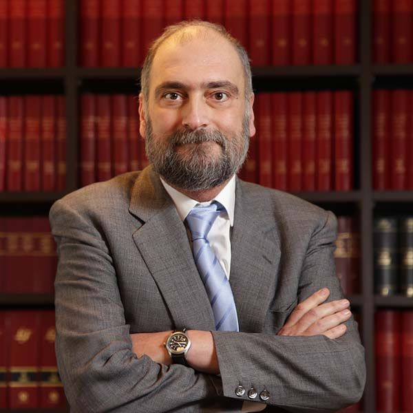 Retrato de Antonio Durán con traje y corbata, de brazos cruzados, posando frente a una estantería llena de libros.